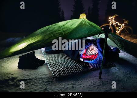 Uomo Barbuto in tenda nel sacco a pelo è guardando al suo telefono a notte d'inverno. Noleggio decorate con luci di Natale a sfondo. Bikepacking e Ch Foto Stock