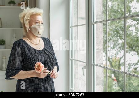 Donna anziana che indossa maschera protettiva e abito nero guardando la finestra a casa Foto Stock