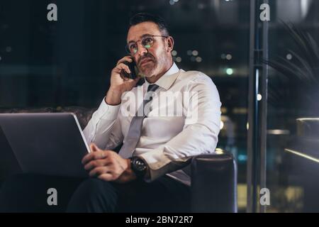 Uomo d'affari che effettua una chiamata mentre aspetta alla lounge dell'aeroporto. Uomo in attesa del suo volo al terminal dell'aeroporto e parlare sul cellulare guardando worri Foto Stock