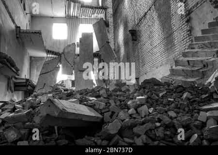 Immagine in bianco e nero. Edificio in cemento distrutto all'interno con un mucchio di detriti di costruzione in primo piano e grandi lastre di cemento appese a reba Foto Stock