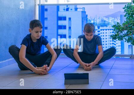 Bambini con computer tablet che svolgono esercizi sportivi, pratica yoga sul balcone. Sport, stile di vita sano, tempo libero attivo a casa Foto Stock