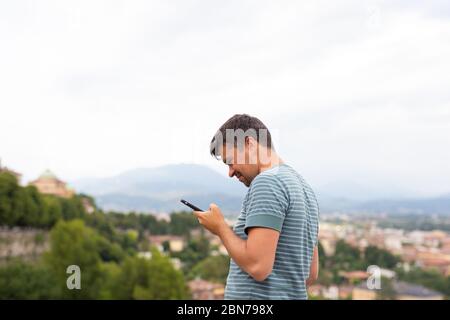 Giovane uomo orologi surf internet tiene sguardi smartphone cellulare in una città. Concetto di viaggio. Viaggiatore. Vacanze estive. Fine della quarantena terminata Foto Stock