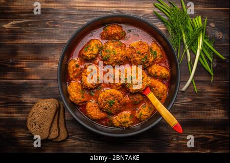Polpette fatte in casa in salsa di pomodoro in una pentola con un cucchiaio di legno. Pane, cipolle verdi e aneto nelle vicinanze. Vista dall'alto Foto Stock