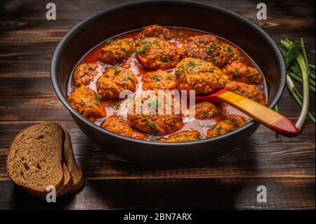 Polpette fatte in casa in salsa di pomodoro in una pentola con un cucchiaio di legno. Pane, cipolle verdi e aneto nelle vicinanze Foto Stock