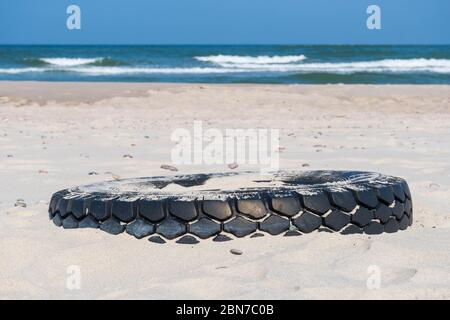 Grande pneumatico nero in gomma lasciato su una spiaggia sabbiosa con mare blu e onde sullo sfondo, ambiente di inquinamento concetto Foto Stock