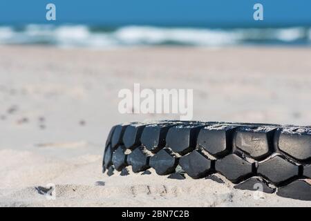 Grande pneumatico nero in gomma lasciato su una spiaggia sabbiosa con mare blu e onde sullo sfondo, ambiente di inquinamento concetto Foto Stock