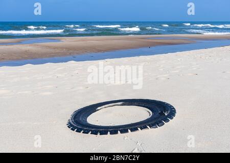 Grande pneumatico nero in gomma lasciato su una spiaggia sabbiosa con mare blu e onde sullo sfondo, ambiente di inquinamento concetto Foto Stock