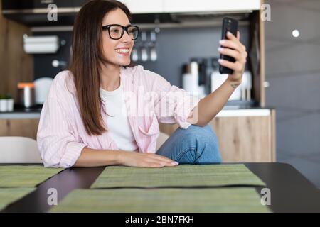 Donna sorridente seduta sul divano della cucina che parla da videocall datazione online guardando il telefono. Video blogger vlogger registrazione vlog a casa. Stile di vita vlo Foto Stock