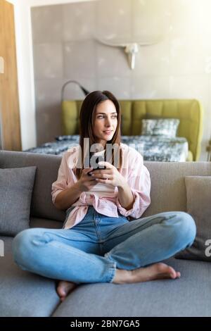 Donna felice e piacevole, millenaria, che si rilassa sul comodo divano, tenendo lo smartphone nelle mani. Sorridente giovane donna che chiacchiera nei social network, guardando f Foto Stock