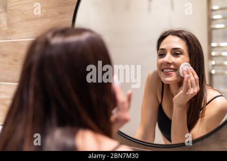 Focalizzato bella giovane donna pulizia la sua pelle con un tampone di cotone, guardando lo specchio nel bagno di casa. Bellezza e cura della pelle concetto, lo stile di vita Foto Stock