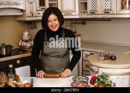 Una donna adulta rotola l'impasto nella sua cucina per preparare la cottura o gnocchi, manti. Foto Stock