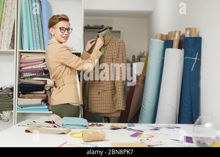 Ritratto di giovane stilista di moda in occhiali in piedi vicino al manichino con giacca e guardando la fotocamera Foto Stock