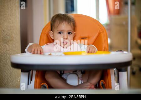 Cute bambina caucasica seduta su una sedia arancione dopo aver mangiato con briciole sul viso, sul tavolo briciole e una piastra di plastica arancione. Chiudere Foto Stock