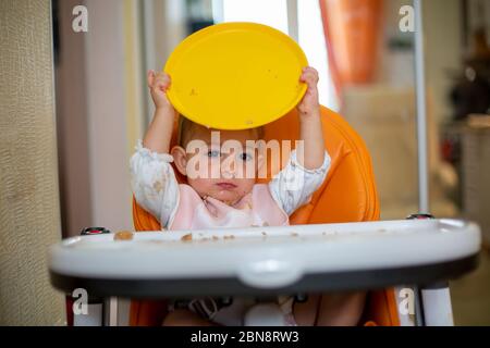 Un bambino caucasico carino si siede su una seggioletta arancione con un tavolo dopo un pasto e gioca con una piastra di plastica arancione. Primo piano, vista frontale, messa a fuoco morbida Foto Stock