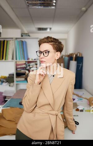 Stilista di moda pensivo in occhiali in piedi e pensando a qualcosa in officina Foto Stock
