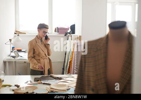Giovane stilista di moda in occhiali che parla sul telefono cellulare che prende ordine mentre lavora in officina Foto Stock