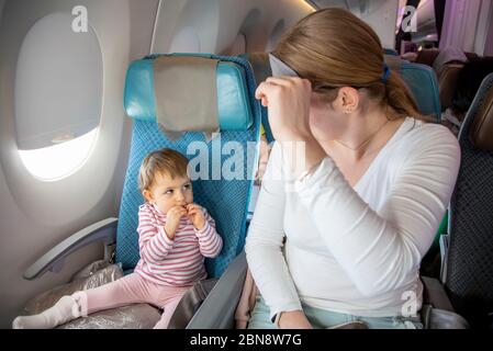 volo comodo con un bambino. carino bimbo e mamma sono seduti in aereo in sedie e si guardano l'un l'altro. mamma solleva una maschera da sonno Foto Stock