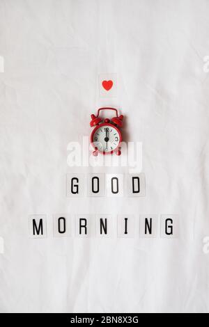Iscrizione Buongiorno, orologio analogico rosso, piccolo cuore su fogli bianchi. Vista dall'alto, disposizione piatta. Verticale. Concetto di riposo, risveglio precoce, sonno Foto Stock