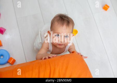 piccolo bambino carino con gli occhi marroni strisciano sull'angolo arancione del letto, cercando di stare in piedi sui piedi e guardando la macchina fotografica, vista dall'alto, primo piano Foto Stock