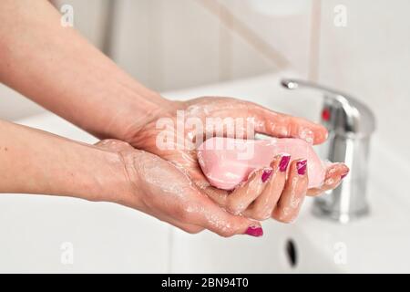 Giovane donna che le lava le mani sotto rubinetto d'acqua con saponetta rosa. Dettaglio sulle dita, unghie ricoperte di pasta viola. Concetto di igiene personale - co Foto Stock