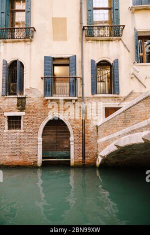 Un edificio in mattoni nelle acque italiane, Venezia. Un ponte di mattoni su un piccolo canale stretto, finestre veneziane classiche nella facciata dell'edificio con Foto Stock