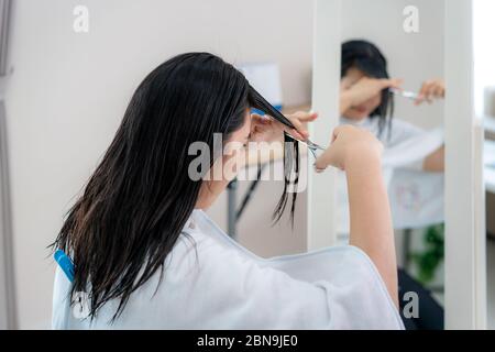 Donna asiatica che taglia i suoi capelli con forbici per taglio di capelli a casa loro rimangono a casa e rifugio in luogo durante il tempo di isolamento casa contro il romanzo Foto Stock
