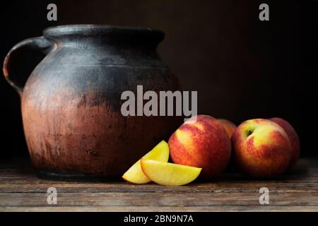 Nettarine fresche, intere e tagliate a fette, accanto ad una caraffa rustica in ceramica, su un tavolo in legno con sfondo scuro Foto Stock