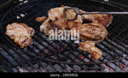 Mano dell'uomo che usa le pinze per girare la carne di pollo sulla griglia per barbecue. Cucina all'aperto. Foto Stock