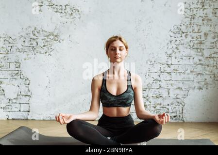 Una bella ragazza giovane è impegnata nella meditazione a casa in una luce interiore in posizione lotus. Cute bionda è impegnata in forma fisica su uno sfondo di un muro di mattoni bianchi. Foto Stock