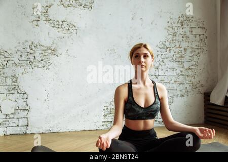 Una bella ragazza giovane è impegnata nella meditazione a casa in una luce interiore in posizione lotus. Cute bionda è impegnata in forma fisica su uno sfondo di un muro di mattoni bianchi. Foto Stock