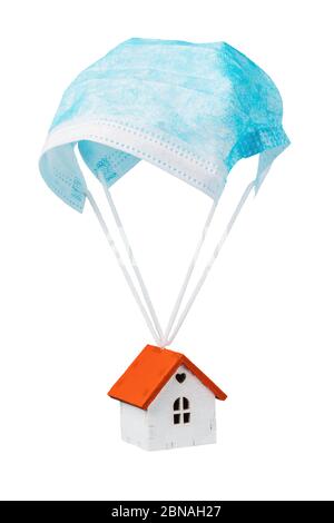 Una casa giocattolo sta volando su un paracadute da una maschera medica, isolata su uno sfondo bianco. Concetto di uscita dalla quarantena Foto Stock