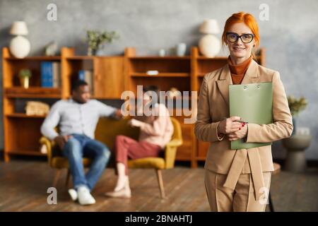 Ritratto di psicologo di successo in piedi con documenti e sorridendo alla fotocamera con coppia seduta sullo sfondo Foto Stock