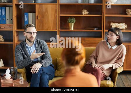 Giovane seduto sul divano con la moglie e parlando con lo psicologo di famiglia in ufficio Foto Stock