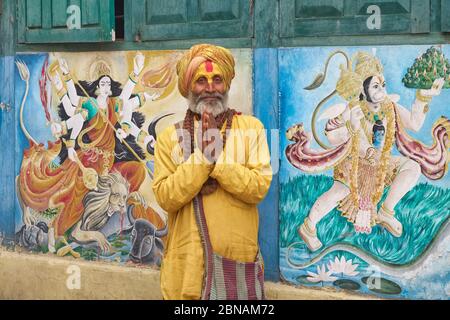 Un sadhu indù (uomo santo) che esegue Namaste (saluto), Tempio di Pashupatinath, Kathmandu, Nepal, tra i murales della dea Durga e il dio delle scimmie Hanuman Foto Stock