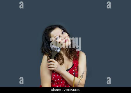 Ritratto di una bella donna di mezza età alla moda con lunghi capelli scuri in un abito rosso, tenendo un pennello e ponendosi su uno sfondo grigio scuro. Foto Stock