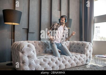 Uomo con la bearded in cuffie inginocchiato sul divano. Foto Stock