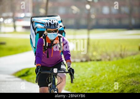 Donna in casco e maschera protettiva con zaino da trasporto grande cibo in bicicletta per strada. Giornata di primavera soleggiata in città. Foto Stock