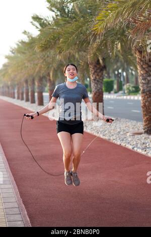 Asian Donna esercizio con una corda di salto con maschera chirurgica protettiva abbassata all'aperto Foto Stock