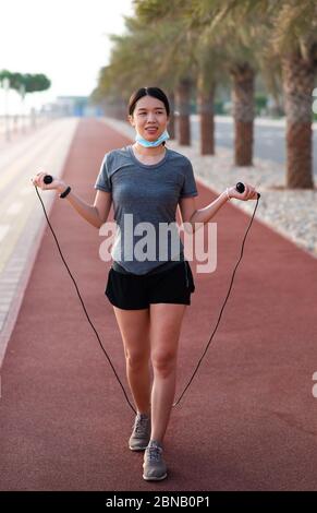 Asian Donna esercizio con una corda di salto con maschera chirurgica protettiva abbassata all'aperto Foto Stock