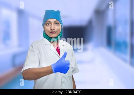 Ritratto di medico lavoratore femminile medico medico indossando maschera chirurgica e cappuccio facendo Thumbs Up, OK segno, eccellente segno. Corridoio esterno dell'ospedale. Covid-19 Foto Stock