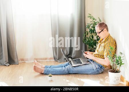 Il giovane è seduto sul pavimento e lavora a casa su un computer con telefono, giornali e una tazza di caffè. Lavoro remoto, lavoro da casa. Foto Stock