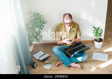 Giovane seduto a terra e lavorando a casa su un computer con telefono, giornali e una tazza di caffè. Lavoro remoto, lavoro da casa. Foto Stock