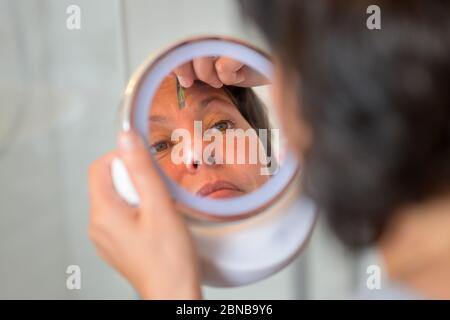 Donna che cazzo le sopracciglia con pinzette usando uno specchio a mano con fuoco al suo riflesso in primo piano Foto Stock