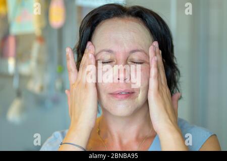 Donna di mezza età che si dona un trattamento di bellezza massaggiando una maschera facciale nella sua pelle con un sorriso di contentment e occhi stretti in un conce di cura della pelle Foto Stock