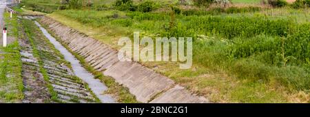 Strada asfaltata con grondaia sul lato della strada o con un fosso per l'acqua e una radura di prato verde lungo la strada. Foto Stock