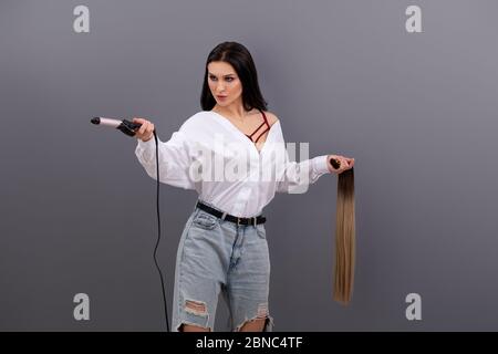 Ritratto di moda di artista o stilista femminile che tiene le sezioni di capelli per l'estensione con il ferro arricciante su sfondo grigio Foto Stock