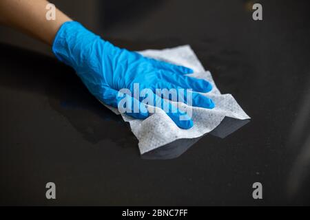 Mano da donna con guanti blu per pulire la superficie del tavolo dell'ufficio domestico con salviette bagnate Foto Stock