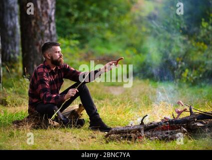 Concetto di andatore affamato. Ragazzo con viso stanco e solitario al picnic o barbecue. Hipster con la barba che cucinano cibo. Uomo, hipster, escursionista arrostire le salsicce sul bastone sul falò in foresta. Foto Stock