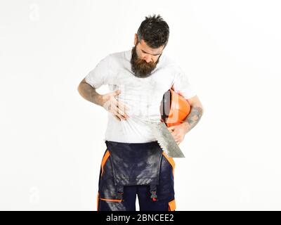 Uomo con barba in abiti sporchi. Costruttore, intonaco, riparatore, caposquadra tiene casco e coltello per stucco, intonacatore, fondo bianco. L'uomo con il volto occupato guarda i vestiti sporchi. Concetto di lavoro polveroso. Foto Stock