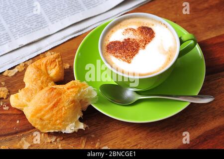 Cappuccino spolverato con cacao a forma di cuore in polvere, accanto a un croissant e un giornale Foto Stock